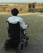 Nancy Mairs sits in her power chair looking at a sign that reads, “No Lifeguard.”